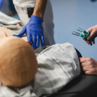 Das Bild zeigt eine Notfall-Simulationspuppe und ein Pflegekraft mit zwei Spritzen in der Hand